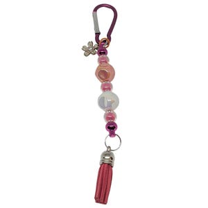 May include: A decorative keychain with a purple carabiner clip. The keychain features a string of pink and iridescent beads, a silver flower charm, and a pink tassel. The metal accents are silver and purple.