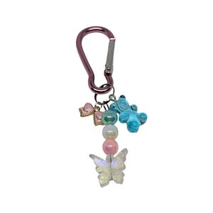 May include: A pink carabiner clip with a collection of charms. Charms include a light blue gummy bear, a pink bow, and a clear butterfly. The butterfly and beads have iridescent qualities.