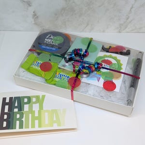 Puede incluir: Una caja de regalo con jabón Dove Men+Care, caramelos, un bolígrafo y una tarjeta de felicitación. La tarjeta dice "HAPPY BIRTHDAY" en verde y negro. La caja está atada con una cinta colorida.