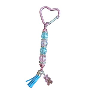 May include: A keychain featuring a pink heart-shaped carabiner attached to a vertical row of alternating light blue and pink crackle glass beads separated by silver-toned rhinestone spacers, ending with a light blue tassel and a small pink bear charm.