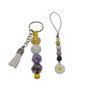 May include: Two beaded accessories: a keychain and a phone charm. The keychain has a silver chain, a white tassel, and a series of yellow, white, purple, and gray beads. The phone charm features similar beads and a white daisy charm.