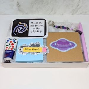Può includere: Una confezione regalo a tema spaziale, contenente un tubetto di crema per le mani, un blocco note con un'etichetta a forma di matita, una carta con un adesivo a forma di pianeta, un portachiavi, una penna e una carta con la scritta "You're the best teacher in the GALAXY!"