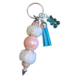 May include: A keychain featuring a central iridescent pink bead flanked by two white faux fur pom-poms and rhinestone-encrusted spacers. It includes a bright blue faux leather tassel and a small translucent blue gummy bear charm on a silver ring.