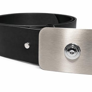 May include: A black belt featuring a rectangular, brushed silver-tone metal buckle. The buckle has rounded corners and a centred, circular, polished chrome-finish accent piece. A single silver-tone metal stud is visible on the black strap.