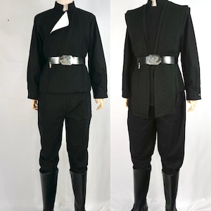 Puede incluir: Dos trajes negros exhibidos en maniquíes. El primero presenta una camisa de manga larga con un acento blanco, pantalones negros, un cinturón plateado y botas negras. El segundo tiene un chaleco, pantalones negros, un cinturón plateado y botas negras.