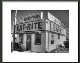 May include: A black and white photo of a diner with the sign "Eat-Rite Diner" above the entrance. The diner has a large window and a door with a sign that says "ATM".