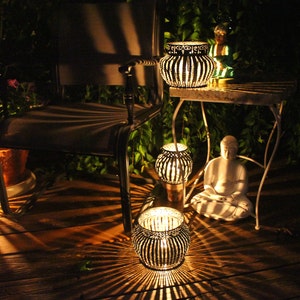 May include: A patio setting with a chair, a table, and several decorative lanterns. The lanterns are made of metal and have a geometric design. The lanterns are lit and casting a warm glow on the surrounding area.