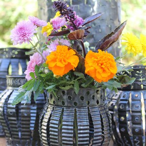 May include: A bouquet of purple, yellow, and orange flowers in a black metal vase with a decorative cut-out design. The vase is made from recycled materials.