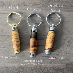 May include: Three keychains with different finishes: gold, chrome, and brushed satin. Each keychain features a wooden handle with a unique wood grain pattern. The handles are labelled "Olive Wood", "Midnight Black Resin & Olive Wood", and "Olive Wood".