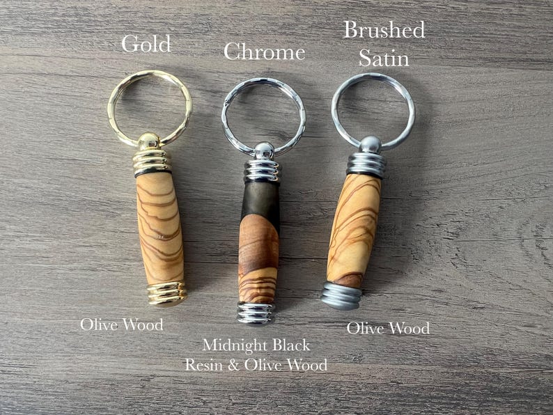 May include: Three keychains with different finishes: gold, chrome, and brushed satin. Each keychain features a wooden handle with a unique wood grain pattern. The handles are labelled "Olive Wood", "Midnight Black Resin & Olive Wood", and "Olive Wood".