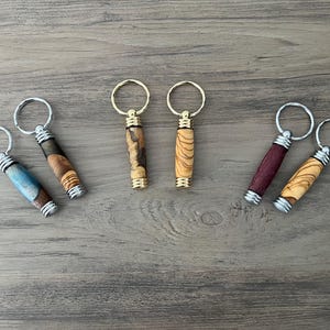 May include: Six wooden keyrings with different wood grain patterns and metal rings. The keyrings are in various colours, including brown, blue, and purple.