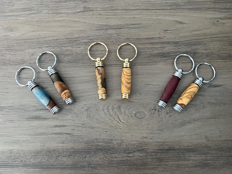 May include: Six wooden keyrings with different wood grain patterns and metal rings. The keyrings are in various colours, including brown, blue, and purple.