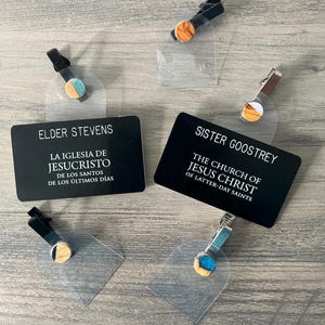 May include: Four name tags with black and silver clips, each with a different wooden circle attached. The circles are made of wood with different colors and patterns. The name tags have white text on a black background. The text on the name tags reads "Elder Stevens", "La Iglesia de Jesucristo de los Santos de los Últimos Días", "Sister Goostrely", and "The Church of Jesus Christ of Latter-day Saints".
