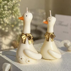 May include: Two off-white duck-shaped candles with orange beaks and gold-colored bows around their necks. The taller candle features small white flower accents on its side, while the shorter candle has a textured white detail on its head.