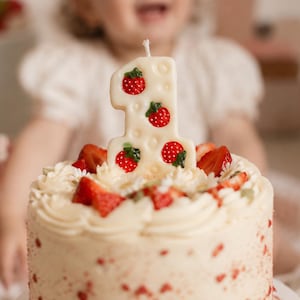 May include: A white number one shaped birthday candle featuring a textured surface with small red strawberry decorations with green leaves, positioned on top of a round cake decorated with white frosting and fresh sliced strawberries.