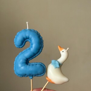May include: A blue number two candle with a textured surface and a white wick, paired with a white goose candle featuring an orange beak and feet, a blue bow around its neck, and a white wick. Both candles are attached to thin wooden sticks.