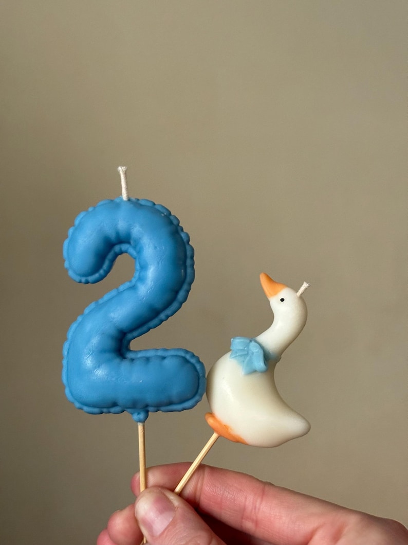 May include: A blue number two candle with a textured surface and a white wick, paired with a white goose candle featuring an orange beak and feet, a blue bow around its neck, and a white wick. Both candles are attached to thin wooden sticks.