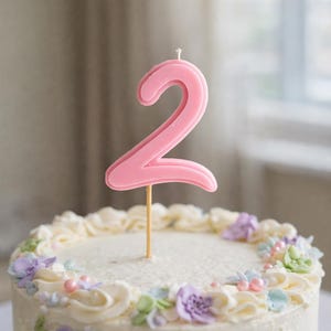 May include: A frosted cake decorated with pastel flowers and a pink number 2 candle. The cake has white frosting with floral accents in shades of purple, blue, and green, and pink pearl sprinkles.