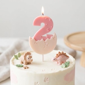 K&ouml;nnte beinhalten: Eine rosa Geburtstagskerze in Form der Zahl zwei, die aus einem Ei schl&uuml;pft, mit brennendem Docht auf einem wei&szlig; glasierten Kuchen. Der Kuchen ist mit kleinen beigen und rosa Dinosaurierfiguren, gr&uuml;nen Bl&auml;ttern und eingepr&auml;gten Fu&szlig;spuren verziert.