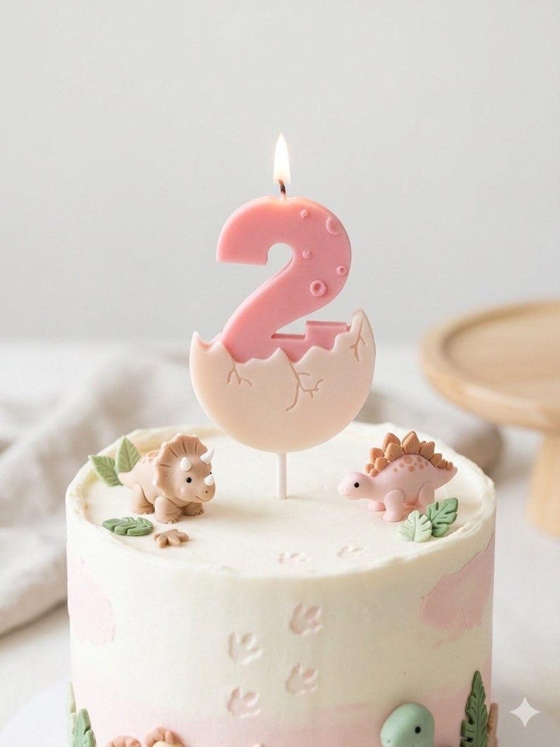 K&ouml;nnte beinhalten: Eine rosa Geburtstagskerze in Form der Zahl zwei, die aus einem Ei schl&uuml;pft, mit brennendem Docht auf einem wei&szlig; glasierten Kuchen. Der Kuchen ist mit kleinen beigen und rosa Dinosaurierfiguren, gr&uuml;nen Bl&auml;ttern und eingepr&auml;gten Fu&szlig;spuren verziert.