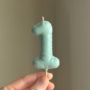 May include: A sage green number one shaped candle with a smooth, matte finish and a stitched-look border detail. The candle features a single white wick at the top and is attached to a thin, pointed wooden stick at the base for insertion.