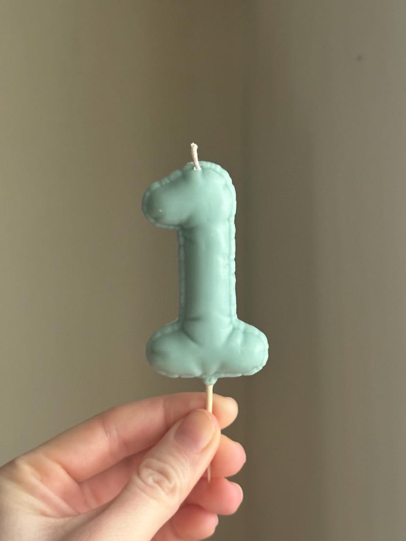 May include: A sage green number one shaped candle with a smooth, matte finish and a stitched-look border detail. The candle features a single white wick at the top and is attached to a thin, pointed wooden stick at the base for insertion.