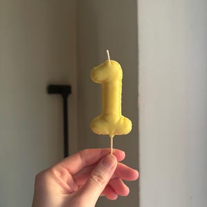 May include: A yellow fabric number one shaped cake topper with a stitched edge and a thin wooden stick extending from the bottom. The soft, padded numeral features a small white wick at the top, mimicking the appearance of a birthday candle.