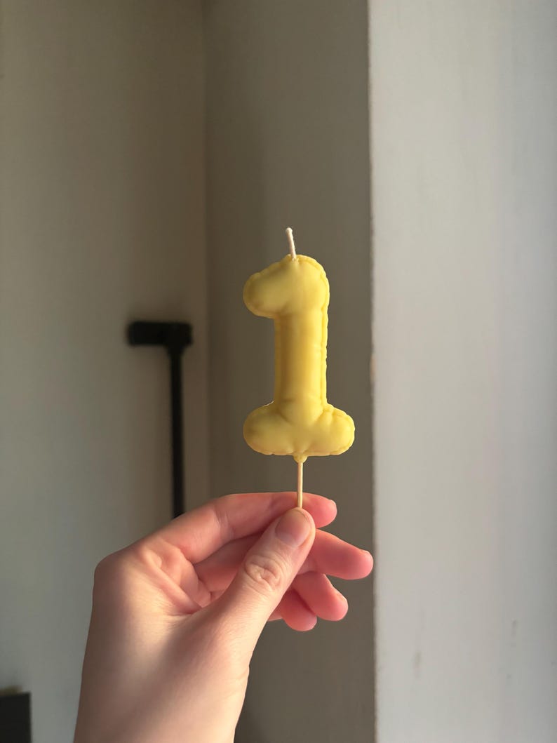 May include: A yellow fabric number one shaped cake topper with a stitched edge and a thin wooden stick extending from the bottom. The soft, padded numeral features a small white wick at the top, mimicking the appearance of a birthday candle.