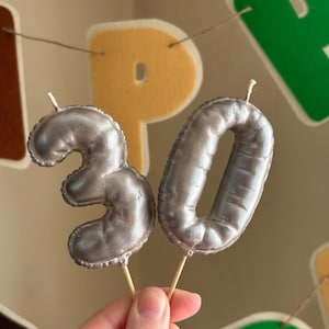 Könnte beinhalten: Silberne Geburtstagskerzen mit der Zahl 30 auf Holzstäben. Die Kerzen sind wie Ballons geformt und werden vor einem Banner mit bunten Buchstaben gehalten. Der Hintergrund ist ein neutrales Beige.