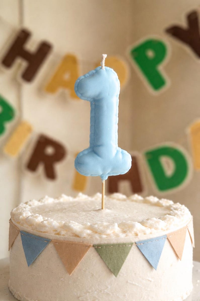 May include: A light blue fabric number one cake topper with a visible stitched edge, mounted on a thin wooden stick. The topper is positioned above a white frosted cake decorated with a small triangular bunting banner in blue, tan, and green.