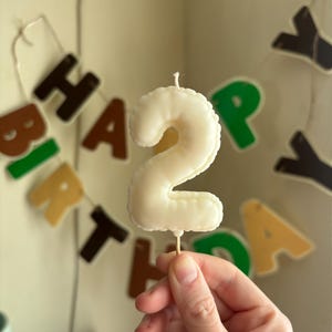 May include: A cream-colored birthday candle shaped like the number two, featuring a smooth, rounded surface and a single white wick at the top. The candle is attached to a thin, light-colored wooden pick at the base for insertion into a cake.
