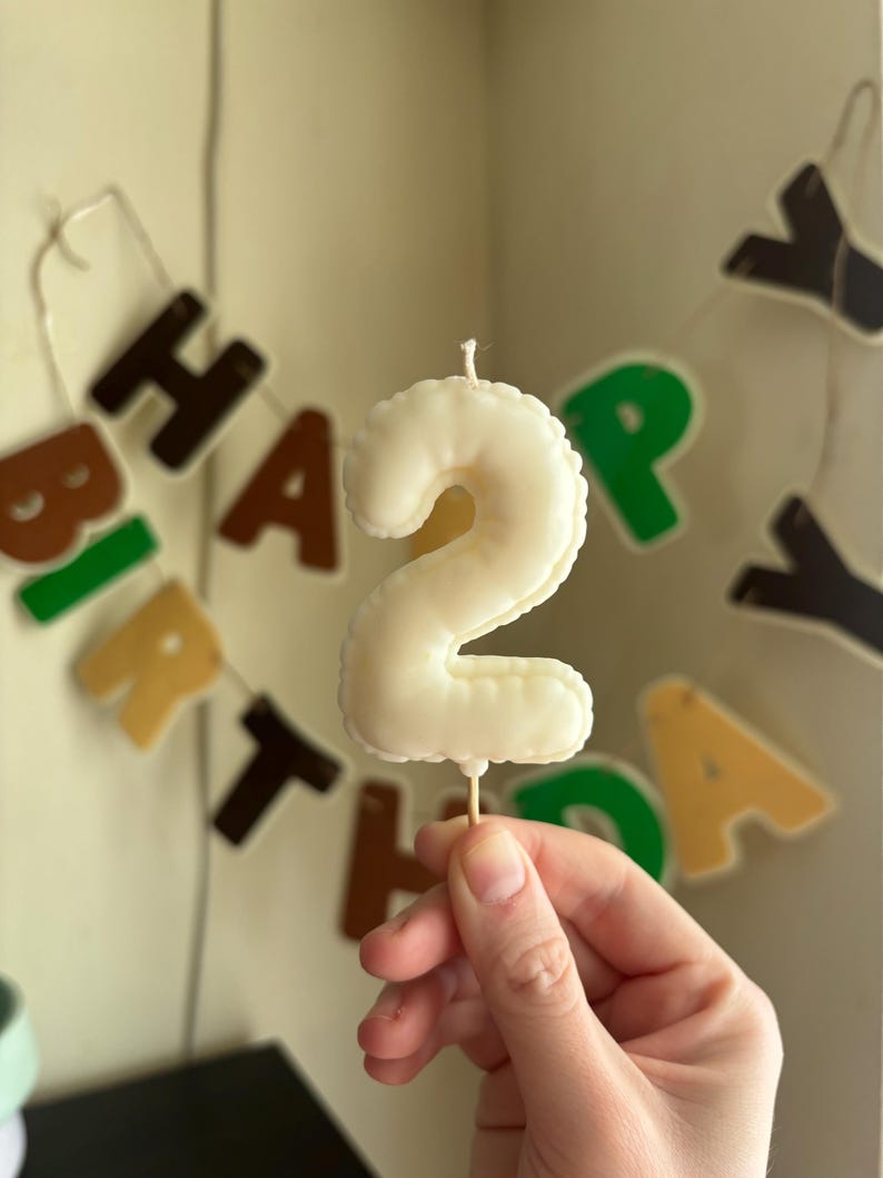 May include: A cream-colored birthday candle shaped like the number two, featuring a smooth, rounded surface and a single white wick at the top. The candle is attached to a thin, light-colored wooden pick at the base for insertion into a cake.