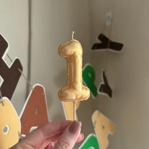 May include: A metallic gold number one candle shaped like a balloon, featuring a smooth, rounded surface and a single white wick at the top. The candle is attached to a thin, straight wooden stick at the base for insertion into a cake or dessert.