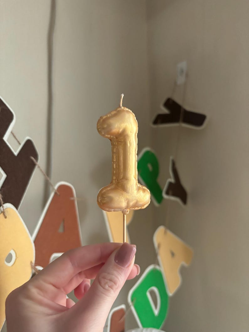May include: A metallic gold number one candle shaped like a balloon, featuring a smooth, rounded surface and a single white wick at the top. The candle is attached to a thin, straight wooden stick at the base for insertion into a cake or dessert.