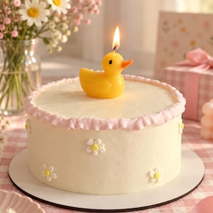 Puede incluir: Un pastel redondo con glaseado blanco liso, decorado con pequeñas flores blancas hechas con manga pastelera con centros amarillos y un borde con volantes rosa claro. Una vela encendida con forma de patito de goma amarillo brillante.