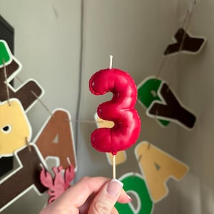 May include: A red number three birthday candle with a glossy finish and a stitched edge detail, mounted on a thin wooden pick. The candle features a single white wick at the top and is shaped like the numeral three with a rounded, puffy appearance.