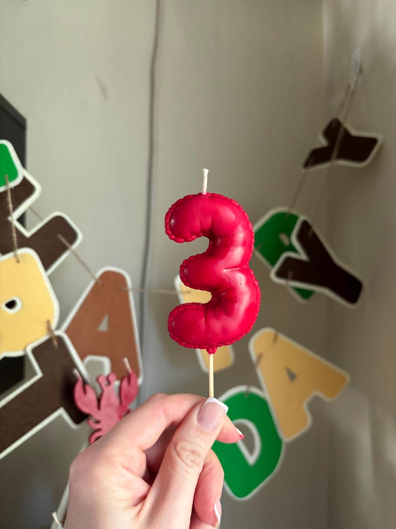 May include: A red number three birthday candle with a glossy finish and a stitched edge detail, mounted on a thin wooden pick. The candle features a single white wick at the top and is shaped like the numeral three with a rounded, puffy appearance.