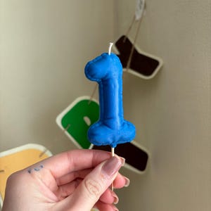 May include: A blue number one shaped candle with a textured surface that mimics the appearance of a balloon. The candle is attached to a thin wooden pick at the base and features a small white wick protruding from the top center of the number.