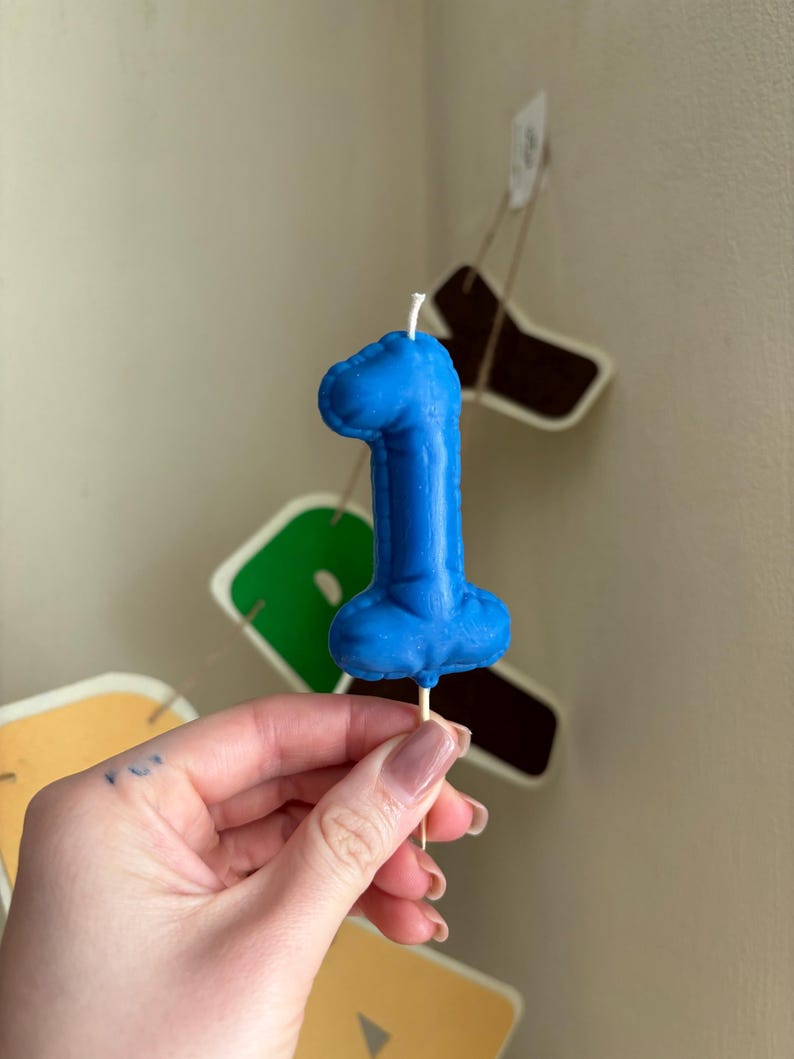 May include: A blue number one shaped candle with a textured surface that mimics the appearance of a balloon. The candle is attached to a thin wooden pick at the base and features a small white wick protruding from the top center of the number.