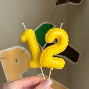 May include: Two bright yellow number candles shaped like the digits one and two. Each candle features a smooth, inflated balloon-like texture and is attached to a thin wooden pick at the base, with a white wick protruding from the top of each number.