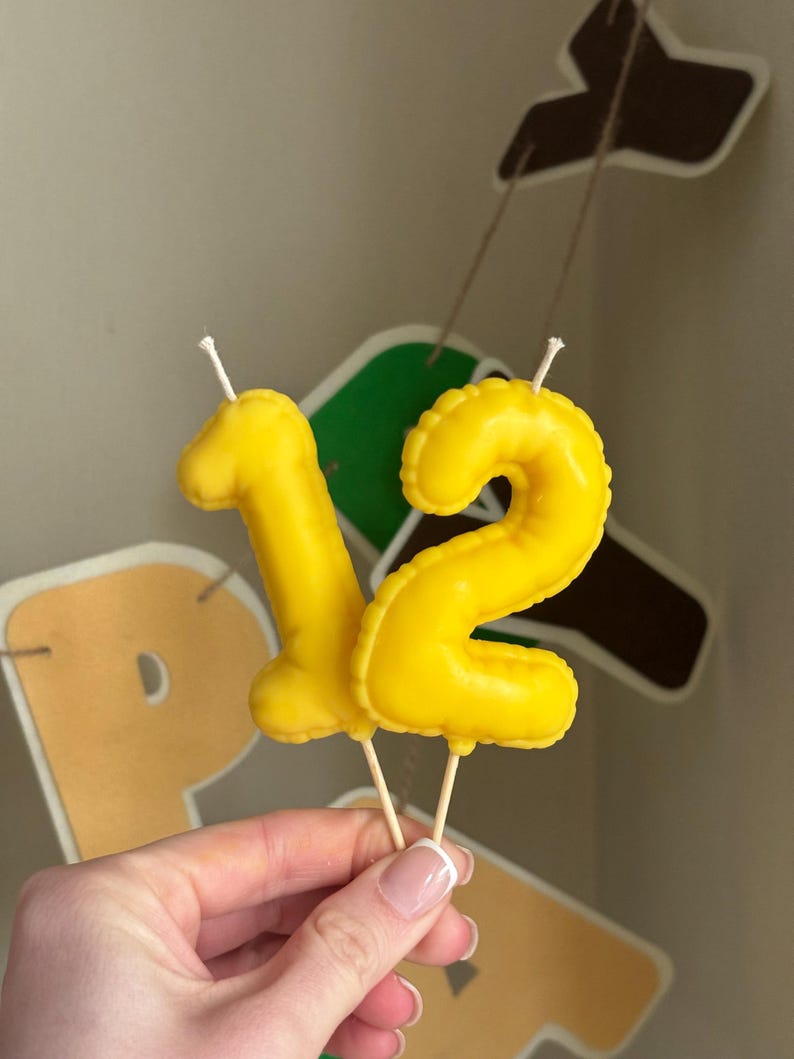 May include: Two bright yellow number candles shaped like the digits one and two. Each candle features a smooth, inflated balloon-like texture and is attached to a thin wooden pick at the base, with a white wick protruding from the top of each number.