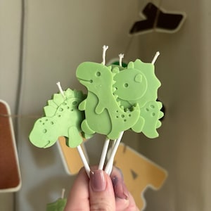 K&ouml;nnte beinhalten: Eine Ansammlung von hellgr&uuml;nen, dinosaurierf&ouml;rmigen Kerzen an wei&szlig;en St&auml;bchen. Jede Kerze zeigt eine eigene Dinosaurier-Silhouette mit gepr&auml;gten Details und einem kleinen wei&szlig;en Docht, der oben aus dem Kopf der Figur herausragt.
