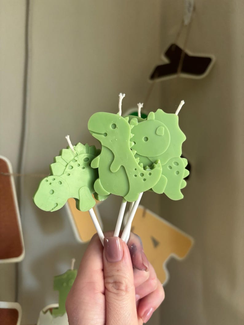 K&ouml;nnte beinhalten: Eine Ansammlung von hellgr&uuml;nen, dinosaurierf&ouml;rmigen Kerzen an wei&szlig;en St&auml;bchen. Jede Kerze zeigt eine eigene Dinosaurier-Silhouette mit gepr&auml;gten Details und einem kleinen wei&szlig;en Docht, der oben aus dem Kopf der Figur herausragt.