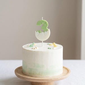 K&ouml;nnte beinhalten: Ein runder, wei&szlig; glasierter Kuchen mit einem hellgr&uuml;nen Aquarellverlauf an der Basis. Die Oberseite ist mit kleinen Dinosaurierfiguren und einer zentralen Kerze in Form der Zahl Drei verziert, die aus einer wei&szlig;en Eierschale ragt.