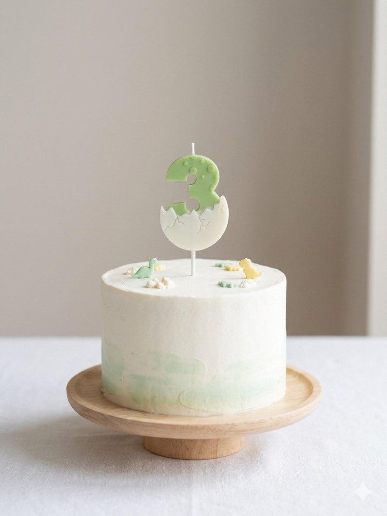 K&ouml;nnte beinhalten: Ein runder, wei&szlig; glasierter Kuchen mit einem hellgr&uuml;nen Aquarellverlauf an der Basis. Die Oberseite ist mit kleinen Dinosaurierfiguren und einer zentralen Kerze in Form der Zahl Drei verziert, die aus einer wei&szlig;en Eierschale ragt.