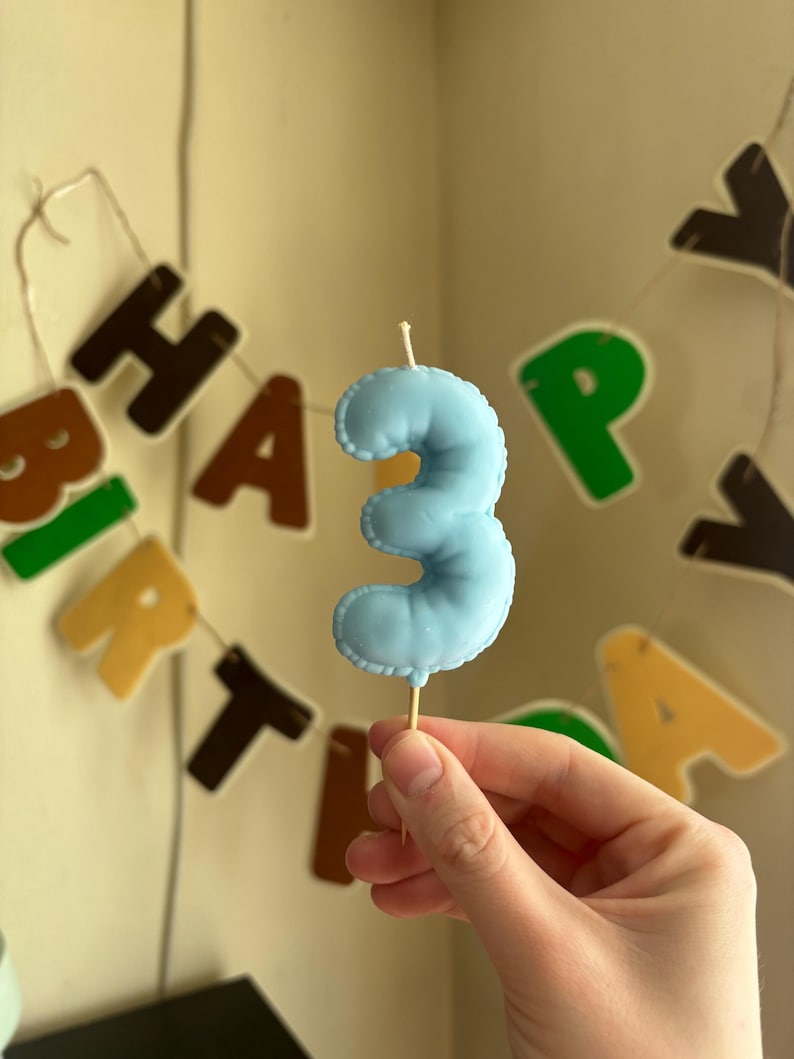 May include: A light blue number three birthday candle with a smooth, rounded surface and a stitched-look border. The candle is mounted on a thin wooden pick, featuring a small white wick at the top, centered against a blurred birthday banner.