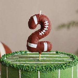 Peut inclure: Gâteau d'anniversaire sur le thème du football américain, orné d'une bougie en forme de chiffre deux. La bougie est marron avec des détails blancs. Le gâteau est décoré avec du glaçage vert et des lignes blanches.