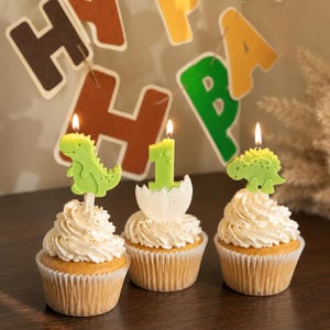K&ouml;nnte beinhalten: Drei Cupcakes mit wei&szlig;em Zuckerguss, jeweils mit einer brennenden Kerze. Eine Kerze ist ein gr&uuml;ner Dinosaurier, eine ist eine gr&uuml;ne Eins in einer wei&szlig;en Eierschale und eine ist ein gr&uuml;ner Stegosaurus. Ein HAPPY BIRTHDAY Banner h&auml;ngt im Hintergrund.