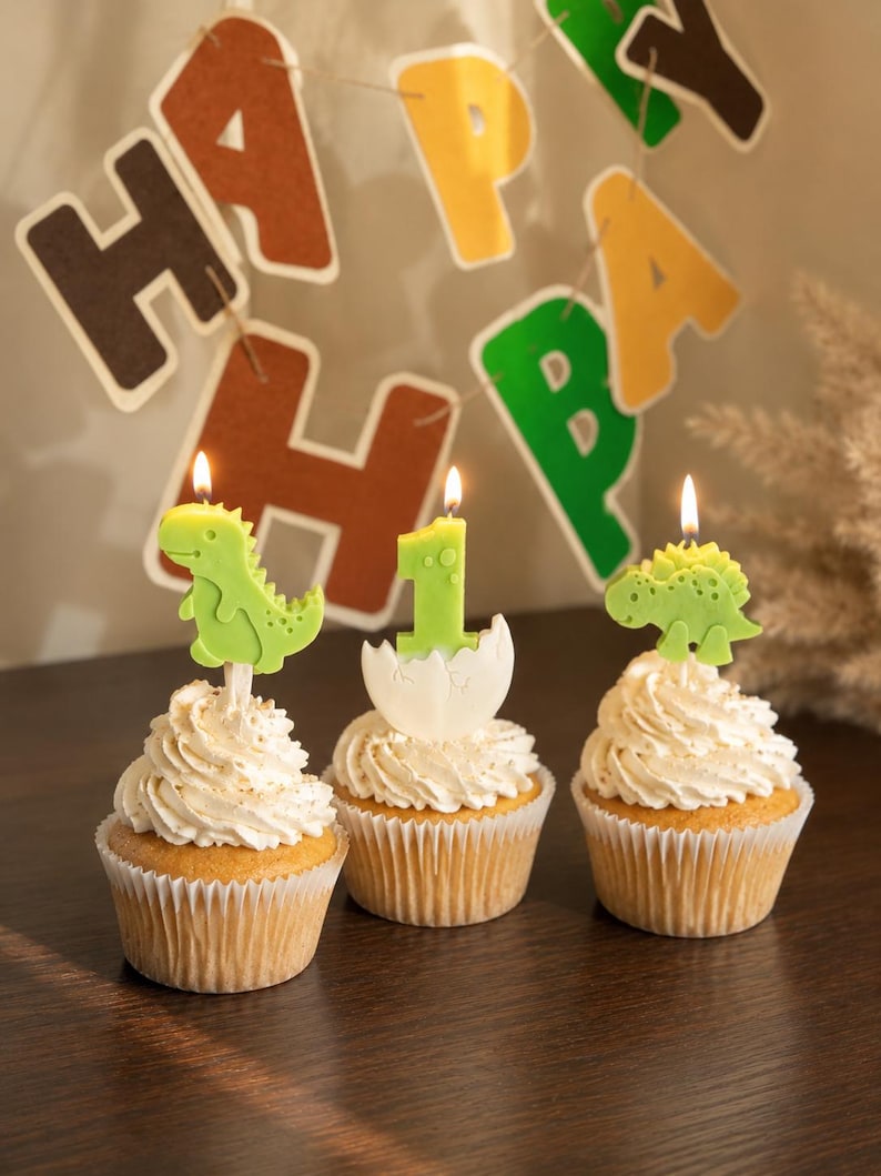 K&ouml;nnte beinhalten: Drei Cupcakes mit wei&szlig;em Zuckerguss, jeweils mit einer brennenden Kerze. Eine Kerze ist ein gr&uuml;ner Dinosaurier, eine ist eine gr&uuml;ne Eins in einer wei&szlig;en Eierschale und eine ist ein gr&uuml;ner Stegosaurus. Ein HAPPY BIRTHDAY Banner h&auml;ngt im Hintergrund.