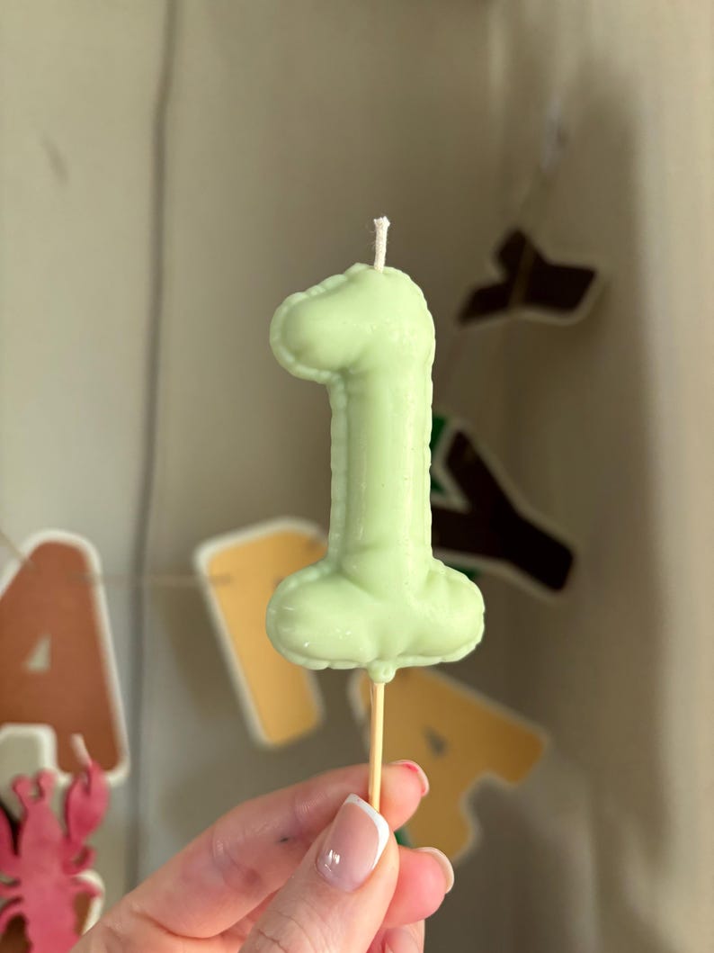 May include: A pale green candle shaped like the number one, featuring a smooth, rounded surface texture that mimics the appearance of a balloon. The candle is mounted on a thin, light-colored wooden stick, with a small white wick at the top.
